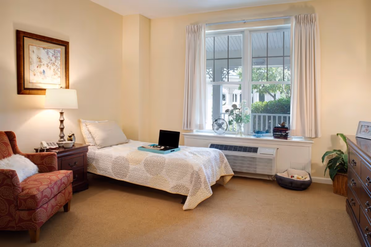 Sunlit bedroom with a single bed, armchair, nightstand and a large window overlooking greenery.