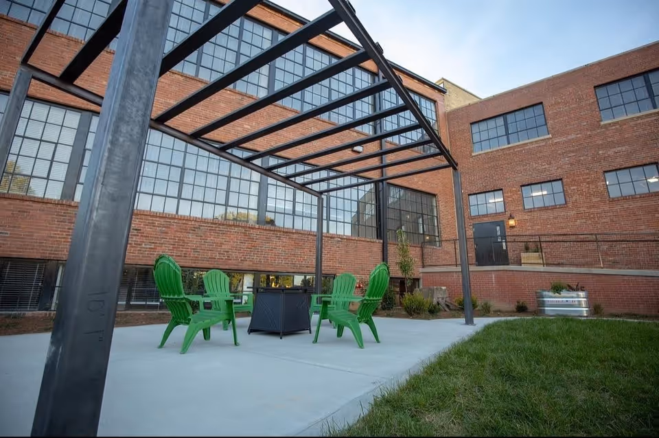 Outdoor patio area with a metal pergola structure, four green Adirondack chairs arranged around a black fire pit, adjacent to a brick building with large industrial-style windows and a grassy area nearby.