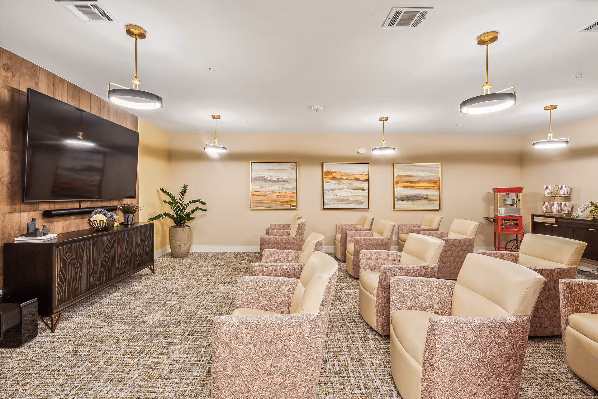 A comfortable media/lounge room with multiple upholstered armchairs arranged facing a large wall-mounted TV, pendant lights, framed artwork, and a popcorn machine.