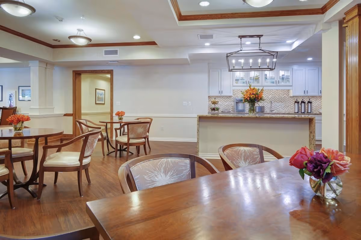 A dining area in a senior living facility with wooden tables and chairs, each table decorated with small flower arrangements. In the background, there is a kitchen counter with a vase of flowers and a modern light fixture hanging above. The room has wooden flooring, white walls with wooden trim, and recessed lighting in the ceiling.