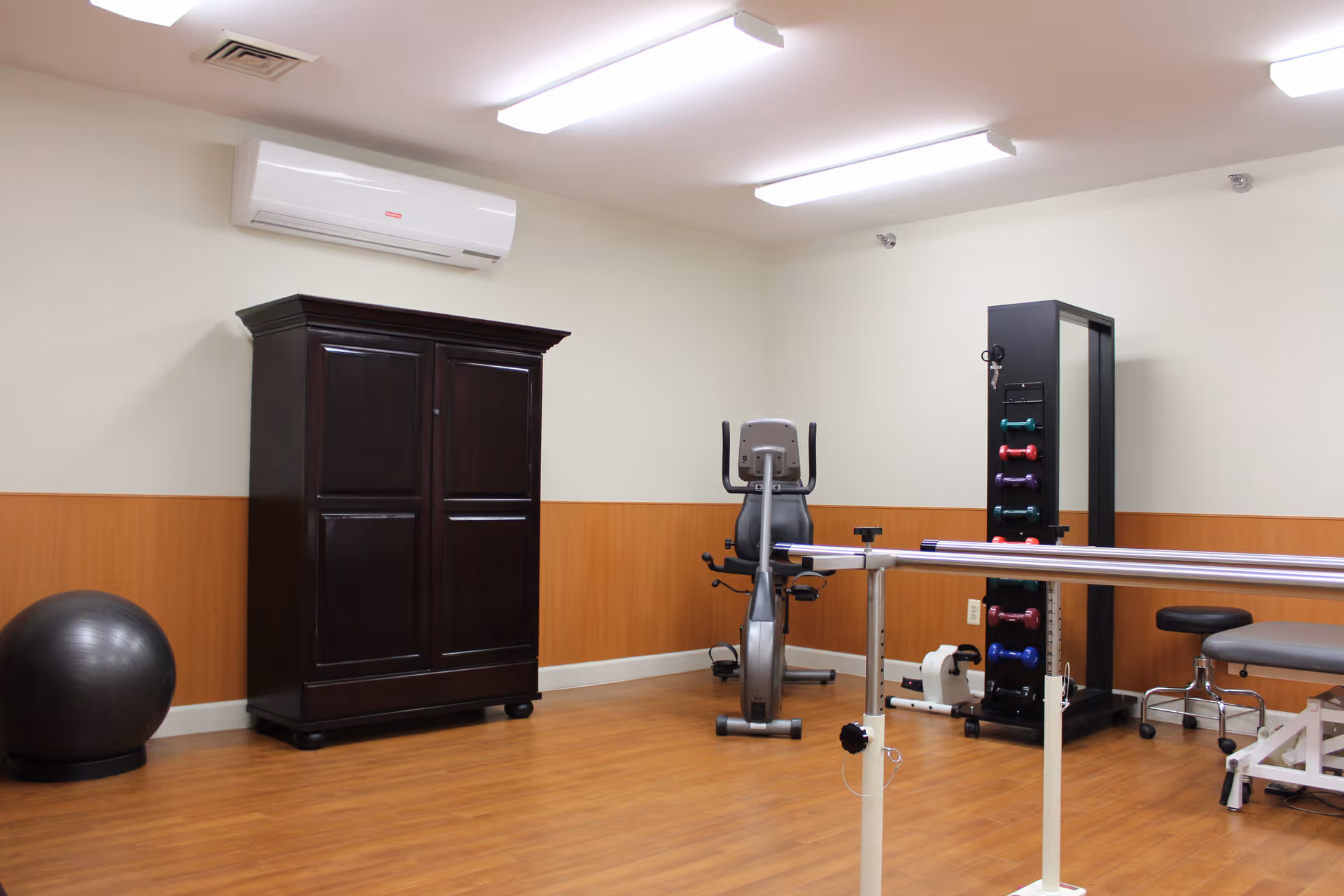 A senior community exercise room with wooden flooring and beige walls. The room contains a black exercise ball, a dark wooden cabinet, a stationary exercise bike, a rack with colorful dumbbells, parallel bars for walking support, and a padded stool. The ceiling has fluorescent lights and an air conditioning unit is mounted on the wall.