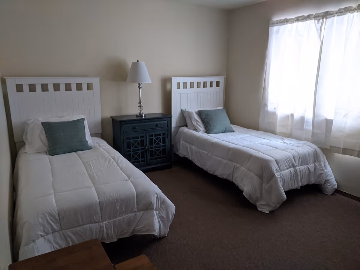 A bedroom with two twin beds, each with white bedding and a green pillow. Between the beds is a dark wooden nightstand with a lamp. A window with sheer white curtains allows natural light into the room.
