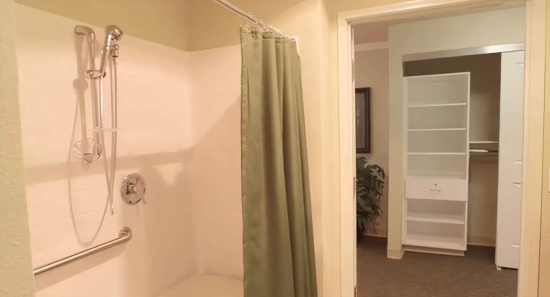 Interior view of a bathroom with a shower area featuring a handheld showerhead, grab bar, and a green shower curtain. Adjacent to the bathroom is an open doorway leading to a room with a white shelving unit and a closet space.