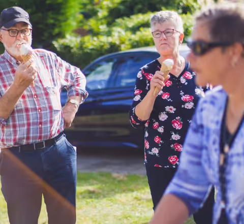 Three elderly people outdoors enjoying ice cream cones. One man wearing a cap and plaid shirt is on the left, a woman with short gray hair and glasses wearing a floral top is in the center, and another woman wearing sunglasses and a blue patterned top is in the foreground on the right. There is a car and greenery in the background.