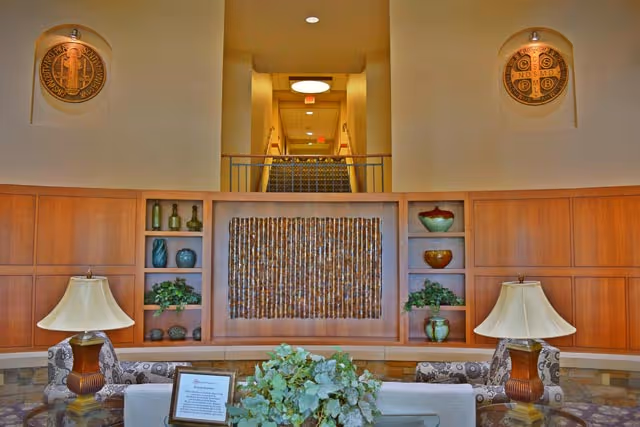 Interior view of a senior living facility lobby area with a seating arrangement including two patterned armchairs and a sofa. There are two table lamps with beige shades on either side of the sofa. Behind the seating area is a wooden wall with built-in shelves displaying decorative vases and plants. Above the shelves is a textured wall art piece. In the background, a carpeted staircase leads to an upper floor, flanked by two circular wall plaques.