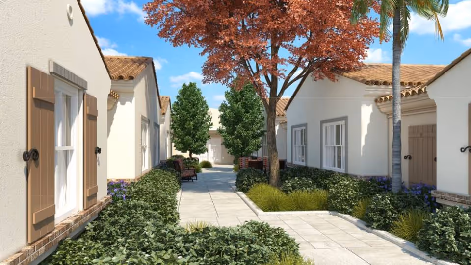 Sunlit courtyard walkway flanked by single-story Mediterranean-style buildings with tiled roofs, landscaping, trees, and a bench.
