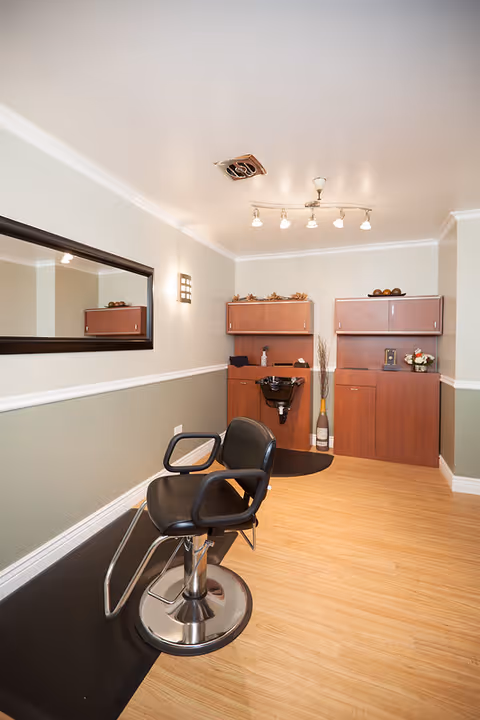 Interior of a salon area in a senior living facility with a black salon chair in front of a black sink, wooden cabinets, a large wall mirror, and light wood flooring.