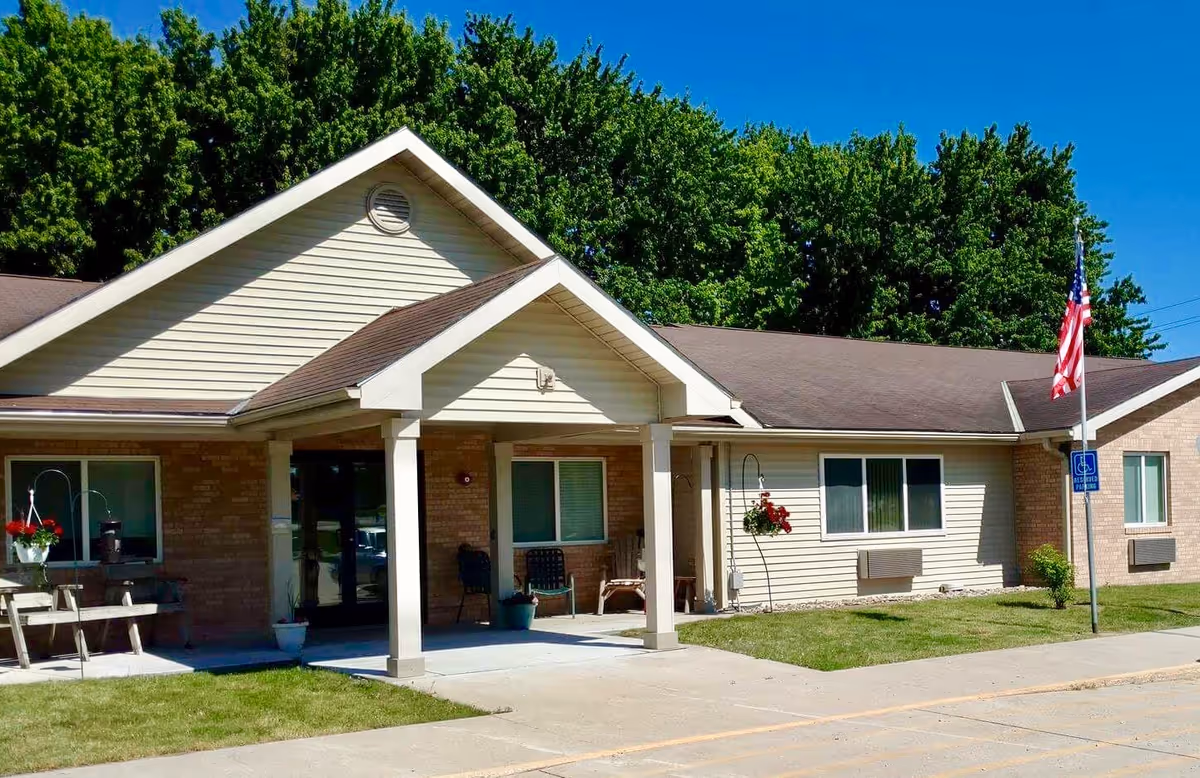 Exterior view of a single-story senior living facility building with beige siding and brick accents, a covered entrance with two columns, a picnic table with hanging flower baskets, an American flag on a pole, and green trees in the background under a clear blue sky.
