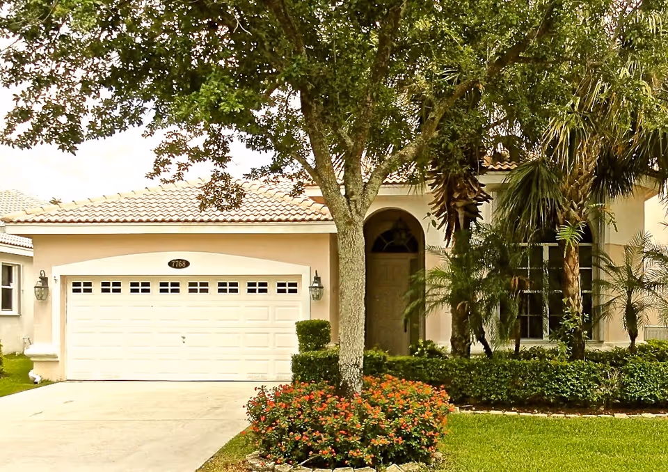 Front exterior view of a single-story house with a tiled roof, a double garage door, a driveway, and a landscaped yard featuring a tree and flowering bushes.