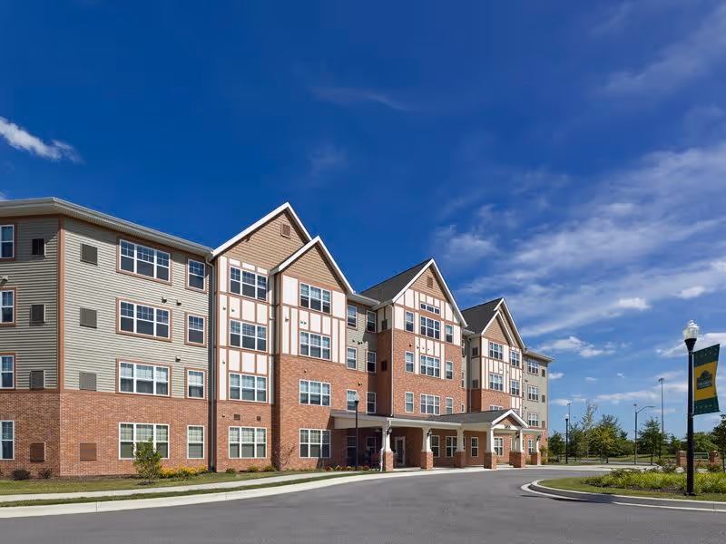 Exterior view of a multi-story senior living facility building with brick and beige siding under a clear blue sky. The building has multiple windows and a covered entrance. There is a driveway and landscaped greenery around the building.