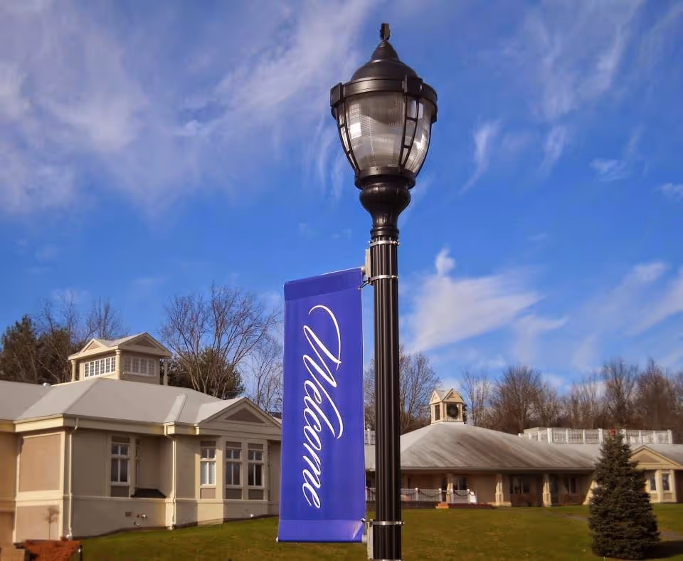 Outdoor view of a senior living facility with a tall black street lamp in the foreground displaying a blue banner that says 'Welcome'. The building in the background has beige walls and a light gray roof, surrounded by trees and a grassy lawn under a partly cloudy blue sky.