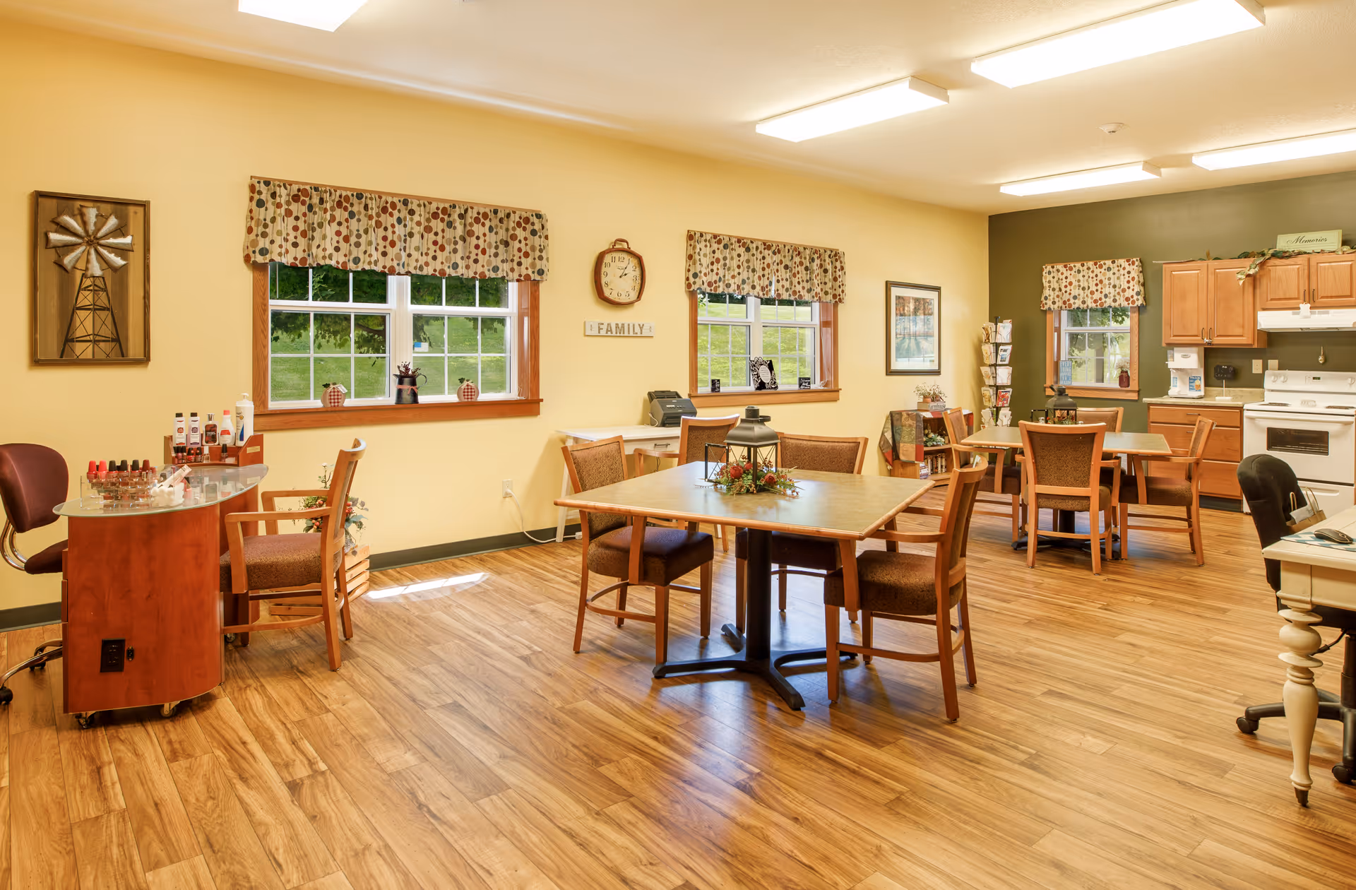 Well-lit common dining and activity room with several tables and chairs, a small kitchenette, and decorative windows and wall art.