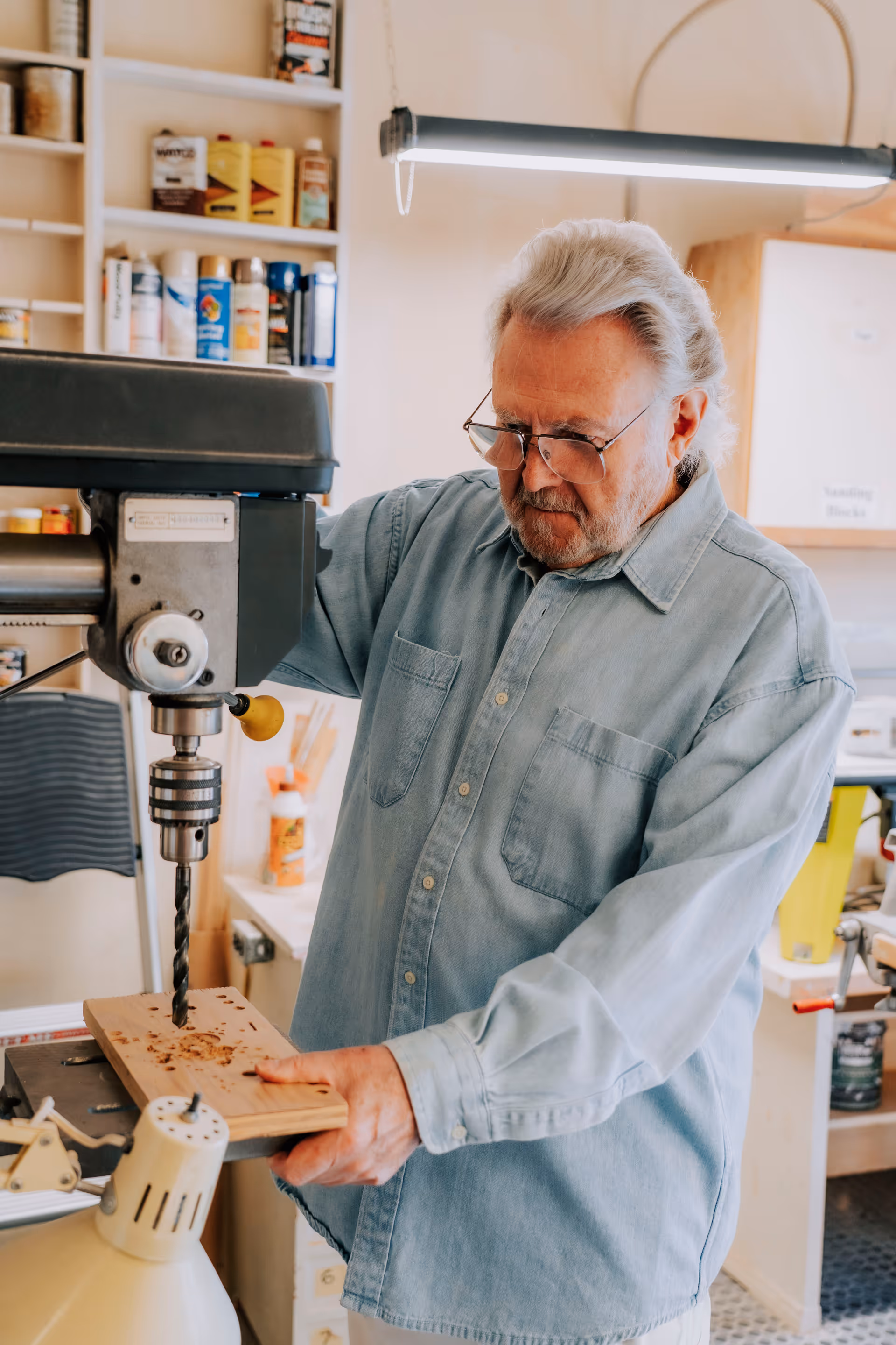 An elderly man wearing glasses and a light blue denim shirt is using a drill press to drill holes into a piece of wood in a workshop setting with shelves of supplies and tools in the background.