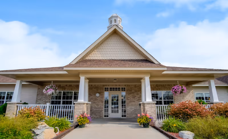 Front exterior view of Stonegate Village Assisted Living & Memory Care building with a covered entrance, brick facade, white pillars, hanging flower baskets, and landscaped bushes and plants along the walkway.