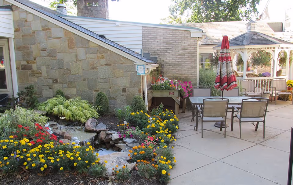Outdoor patio area at Presbyterian Village at Hollidaysburg featuring a stone wall, colorful flower beds with yellow, red, and orange flowers, a small water feature with rocks, a table with four chairs, a closed red and white striped umbrella, and a white gazebo with hanging flower baskets in the background.