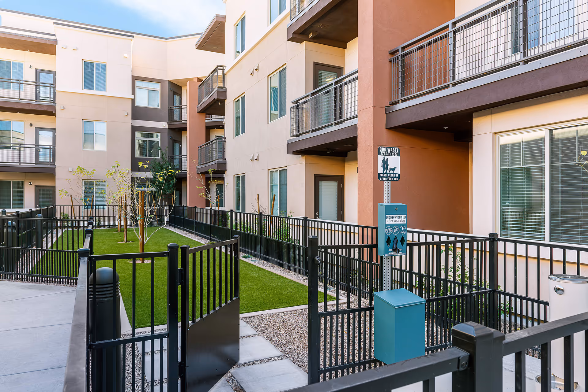 Outdoor fenced dog waste station area with artificial grass and small trees, surrounded by a multi-story residential building with balconies and windows.