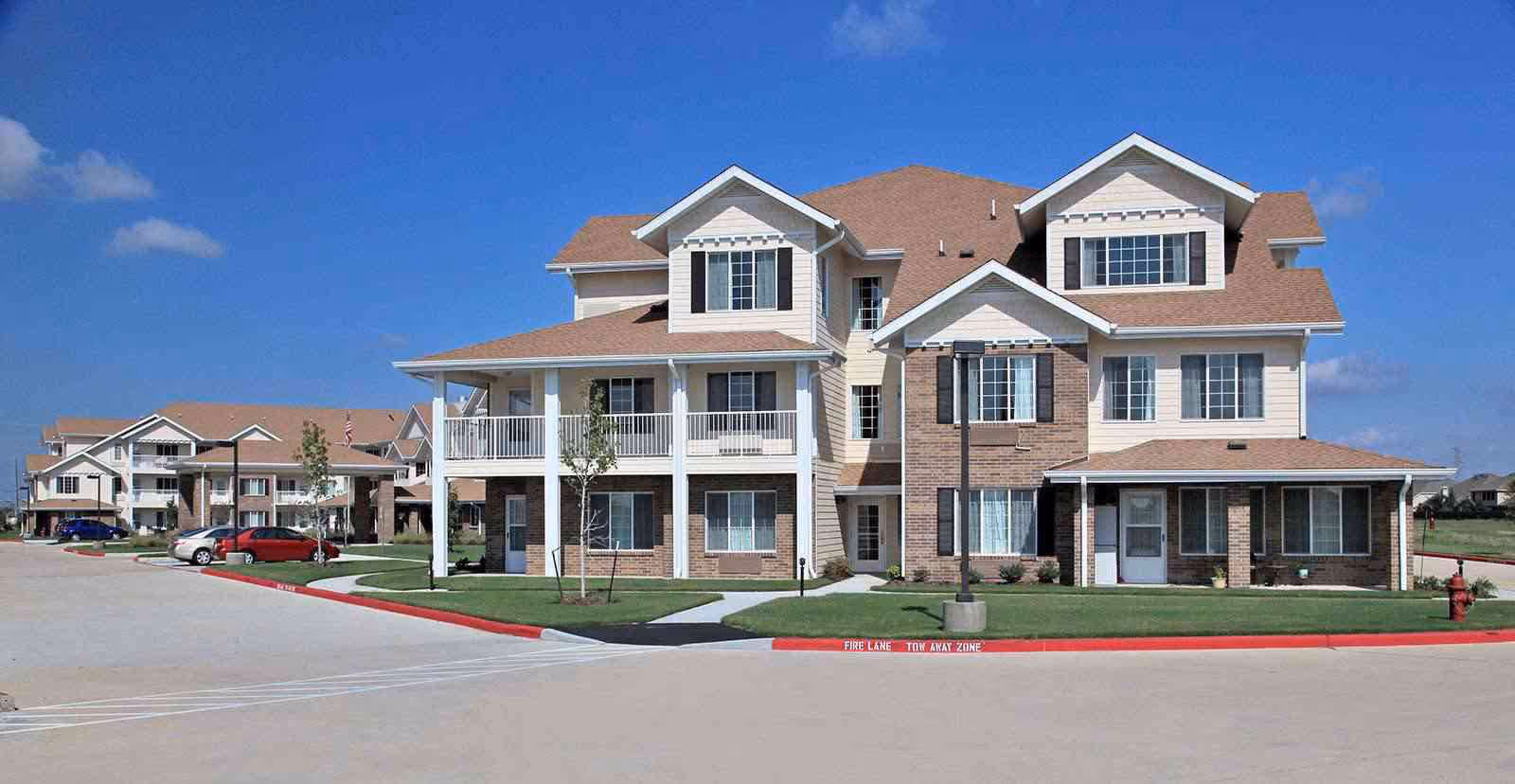 Exterior view of a multi-story residential building with beige siding and brick accents, featuring multiple windows, balconies, and a covered entrance. The building is surrounded by a paved driveway, green lawns, and a few small trees under a clear blue sky.