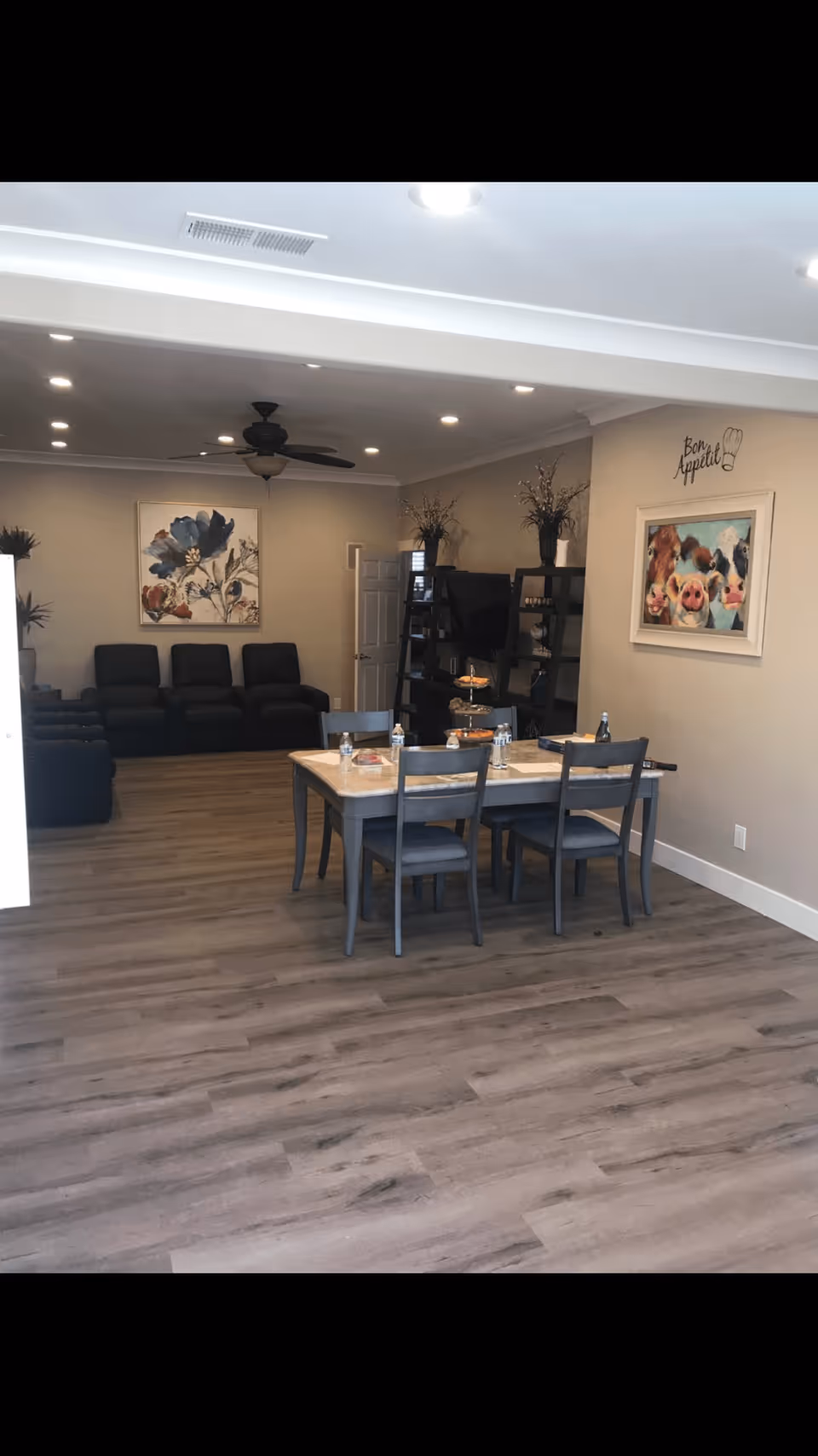 Interior view of a living and dining area with a wooden dining table and four chairs in the foreground. The table has water bottles and snacks on it. In the background, there are three black recliner chairs against the wall, a ceiling fan, and a shelving unit with a TV and decorative items. The walls are beige with framed artwork, including a floral painting and a colorful painting of farm animals with the words 'Bon Appetit' above it. The floor is light wood laminate.