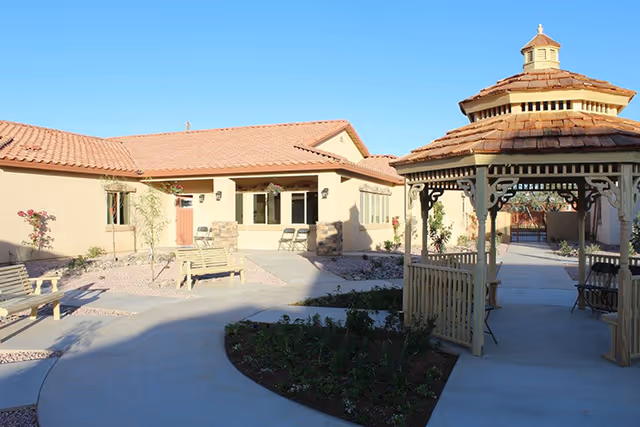 Outdoor courtyard area of a senior care community featuring a wooden gazebo with a shingled roof, benches, small plants, and a paved walkway surrounded by beige buildings with red tile roofs under a clear blue sky.