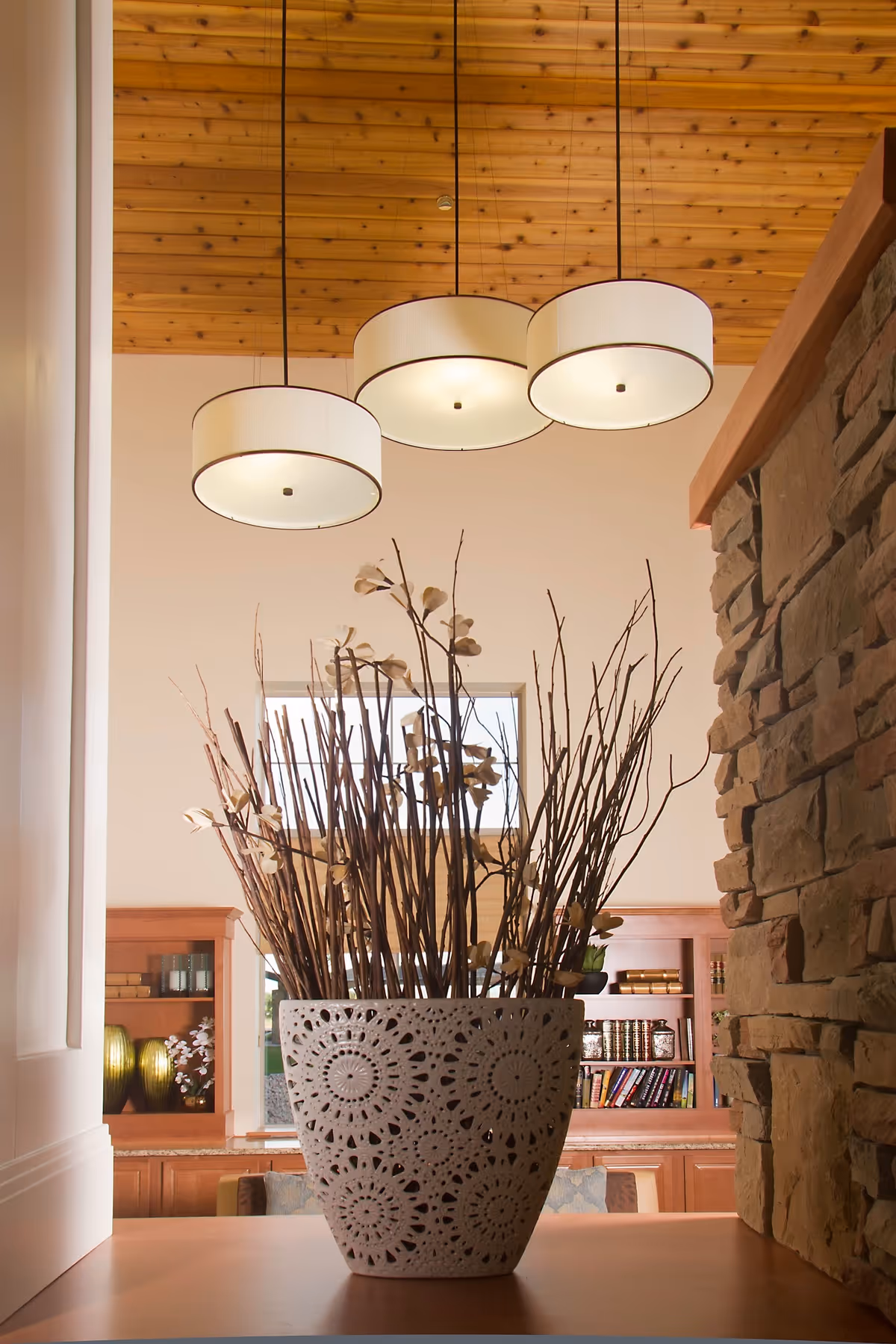 A decorative white ceramic pot with intricate cut-out patterns holding tall dried branches and flowers, placed on a wooden surface. Behind it, there is a stone wall on the right and wooden shelves with books and decorative items in the background. Three modern round pendant lights hang from a wooden ceiling above.