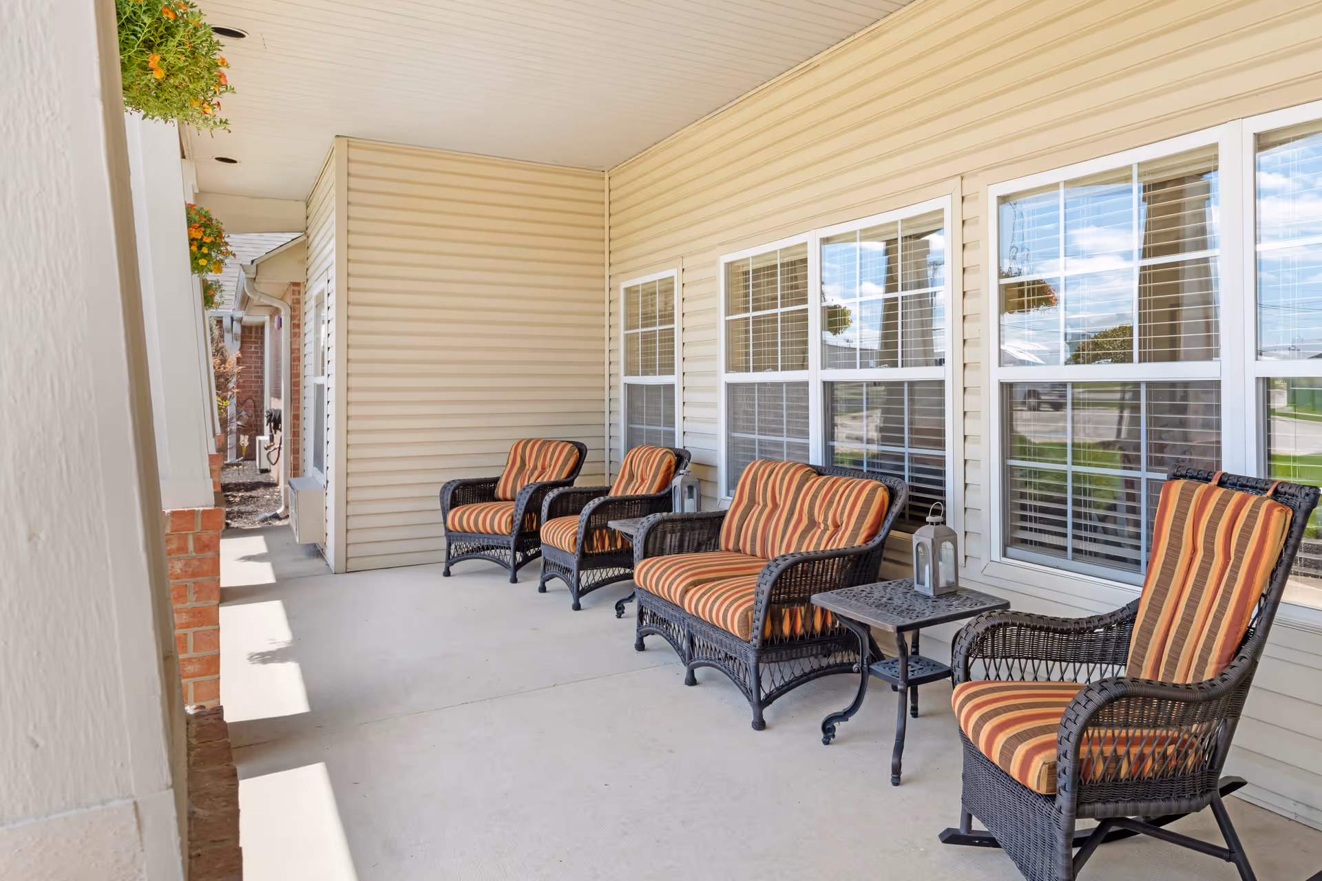 A covered outdoor patio area with four wicker chairs and a loveseat, all with striped orange and brown cushions. Two small black metal tables with lanterns are placed between the seating. The patio is adjacent to a beige siding building with multiple windows and hanging flower baskets.