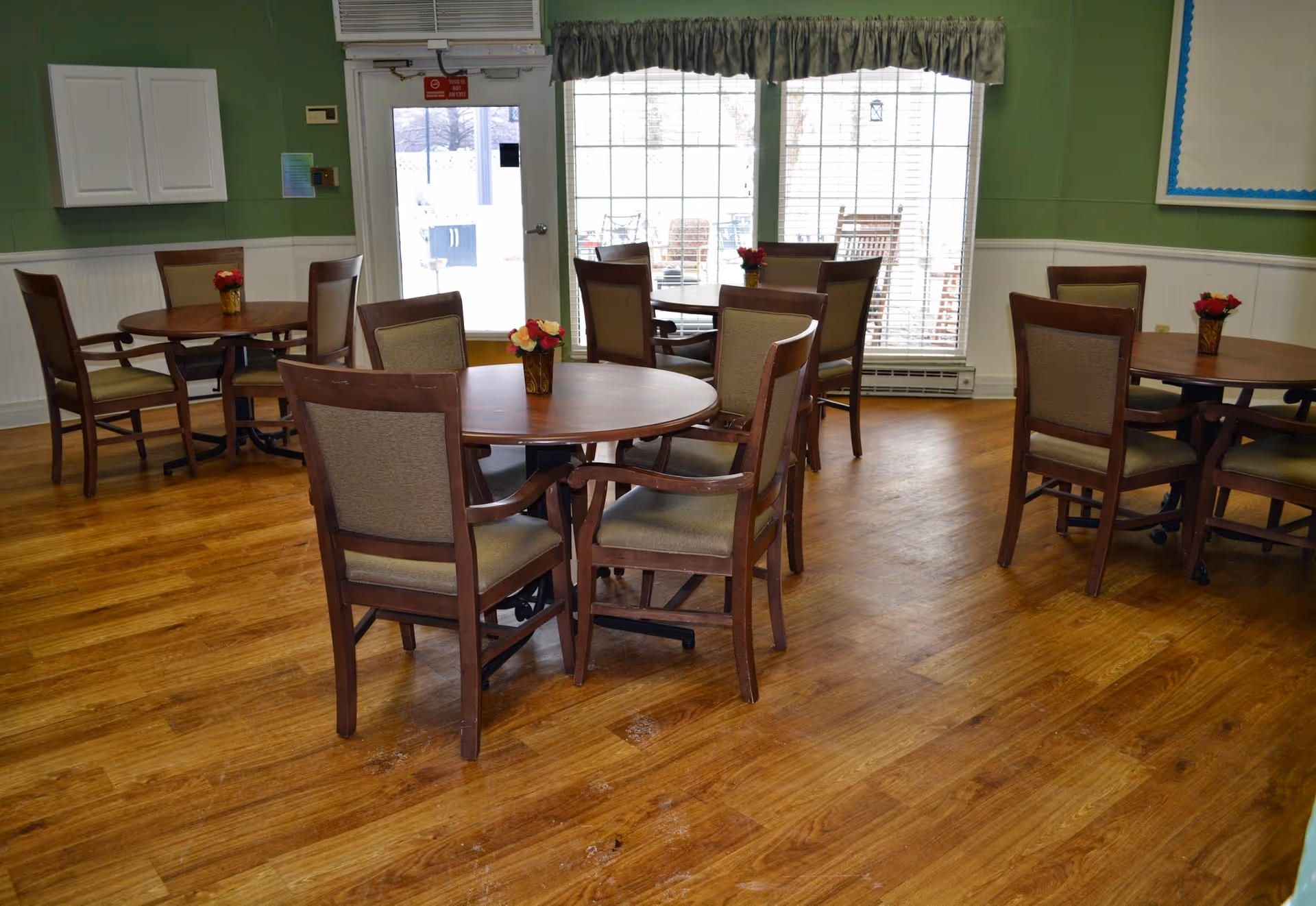 A dining room with several round wooden tables, each surrounded by four cushioned chairs. Small flower arrangements are placed in the center of each table. The room has wooden flooring, green walls with white wainscoting, a door with a window, and large windows covered with curtains letting in natural light.