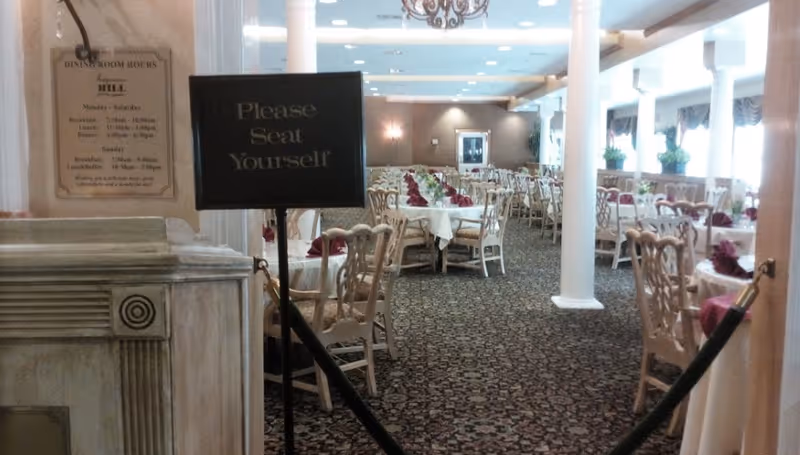 View of a dining room with multiple tables covered with white tablecloths and burgundy napkins, surrounded by wooden chairs. A sign in the foreground reads 'Please Seat Yourself.' The room has carpeted floors, white columns, and large windows with curtains allowing natural light in.