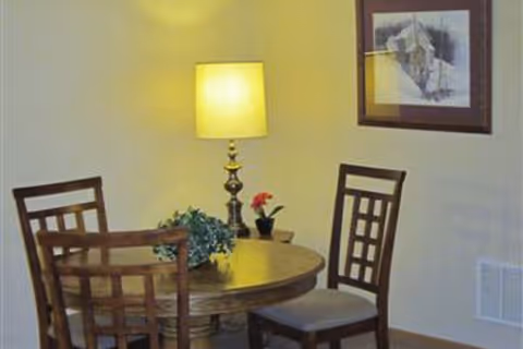 Small dining nook with a round wooden table, four chairs, a lit table lamp, potted plants, and framed artwork on the wall.