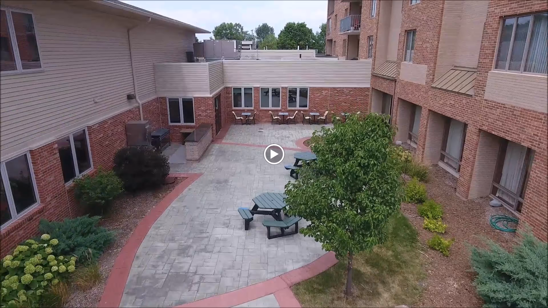 Outdoor courtyard area of a senior living facility with brick buildings surrounding a paved patio. The patio has several green picnic tables with attached benches, a small tree, and some shrubs and plants along the edges. There are also a few chairs and tables near the building walls.