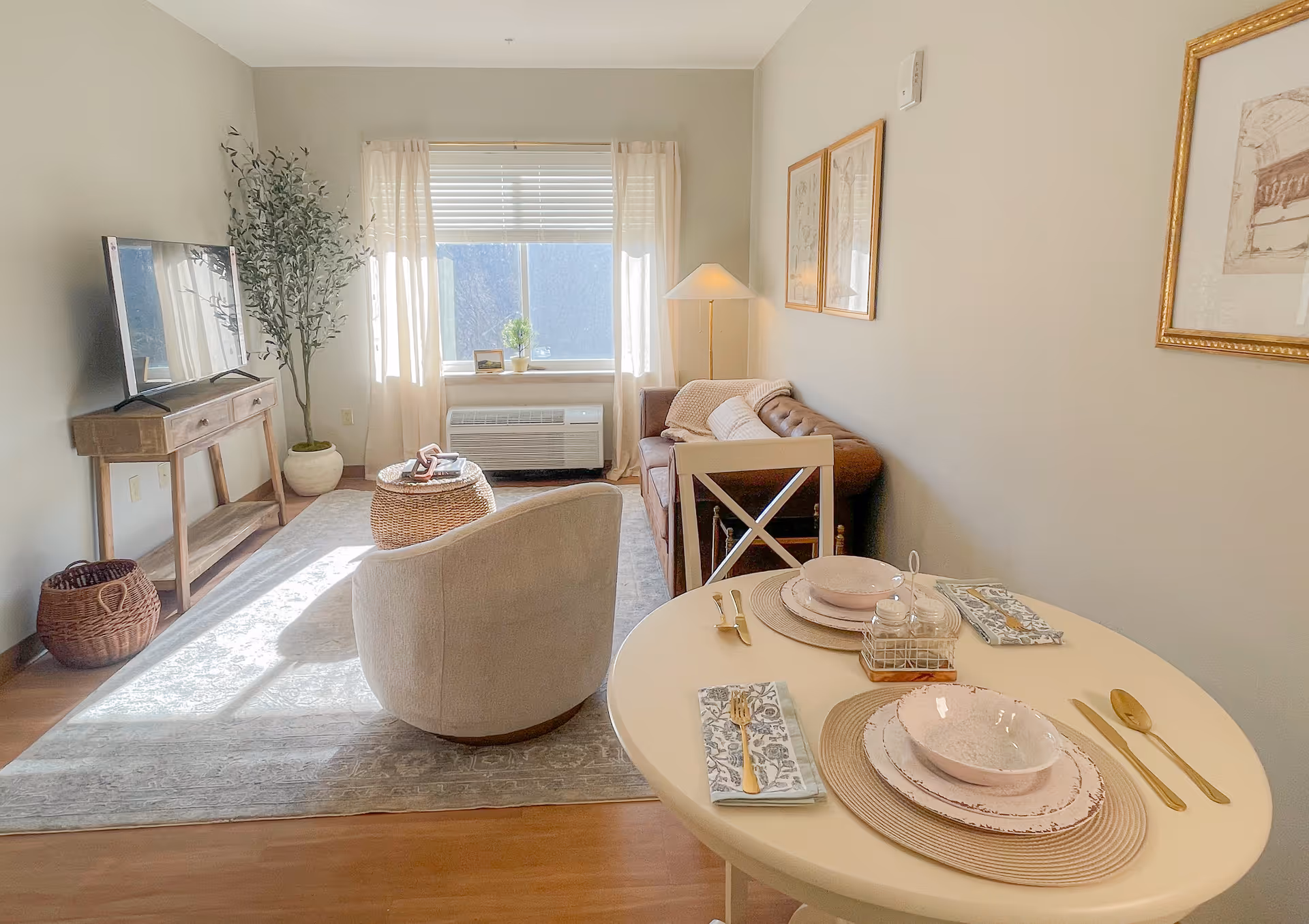 Sunlit living room and dining area with a round set table, an armchair, sofa, TV on a console, and a window with curtains.