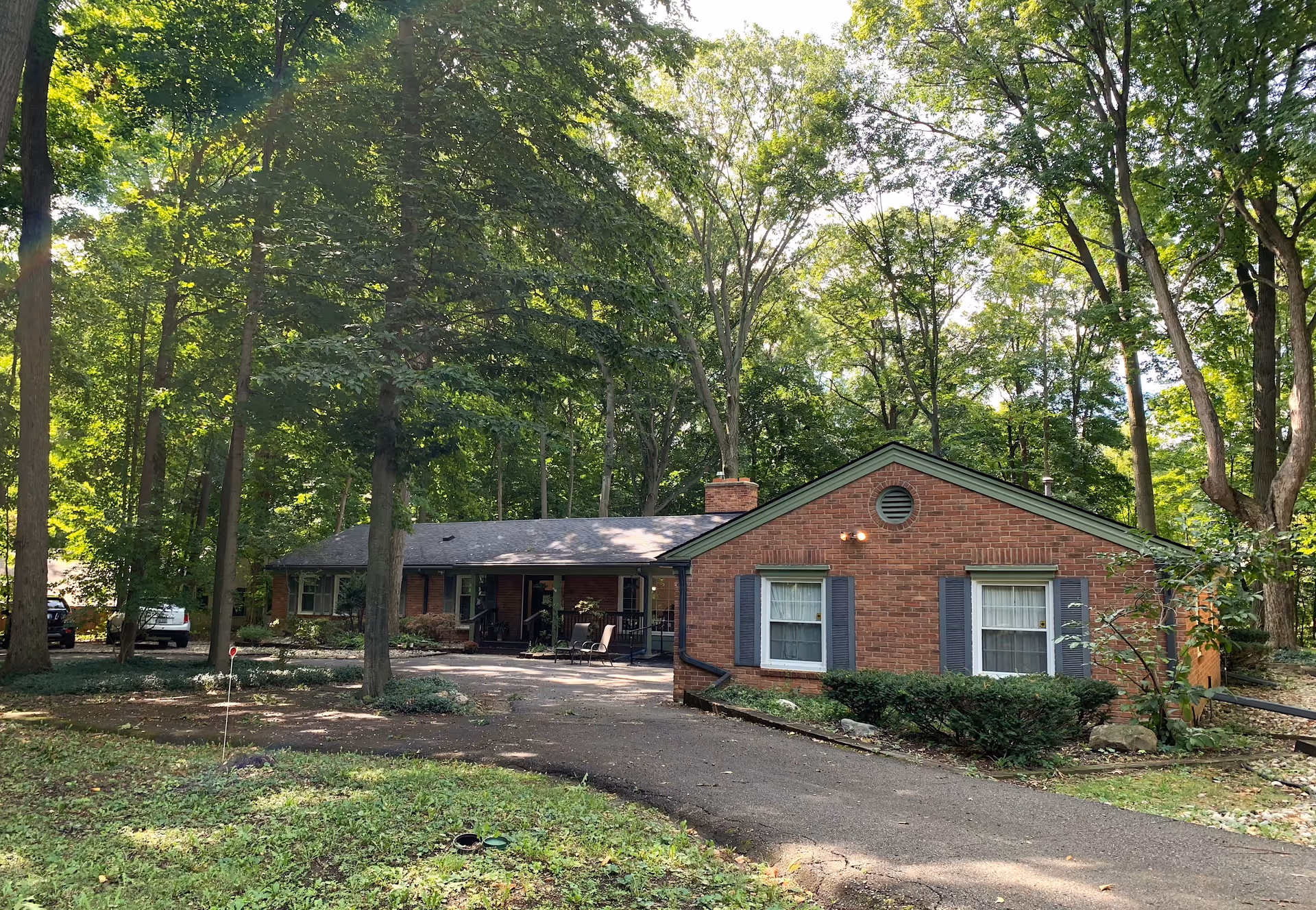 A single-story brick building surrounded by tall trees and greenery, with a paved driveway leading up to the entrance. The building has two windows with blue shutters and a small porch area with chairs. The scene is well-lit with natural sunlight filtering through the trees.