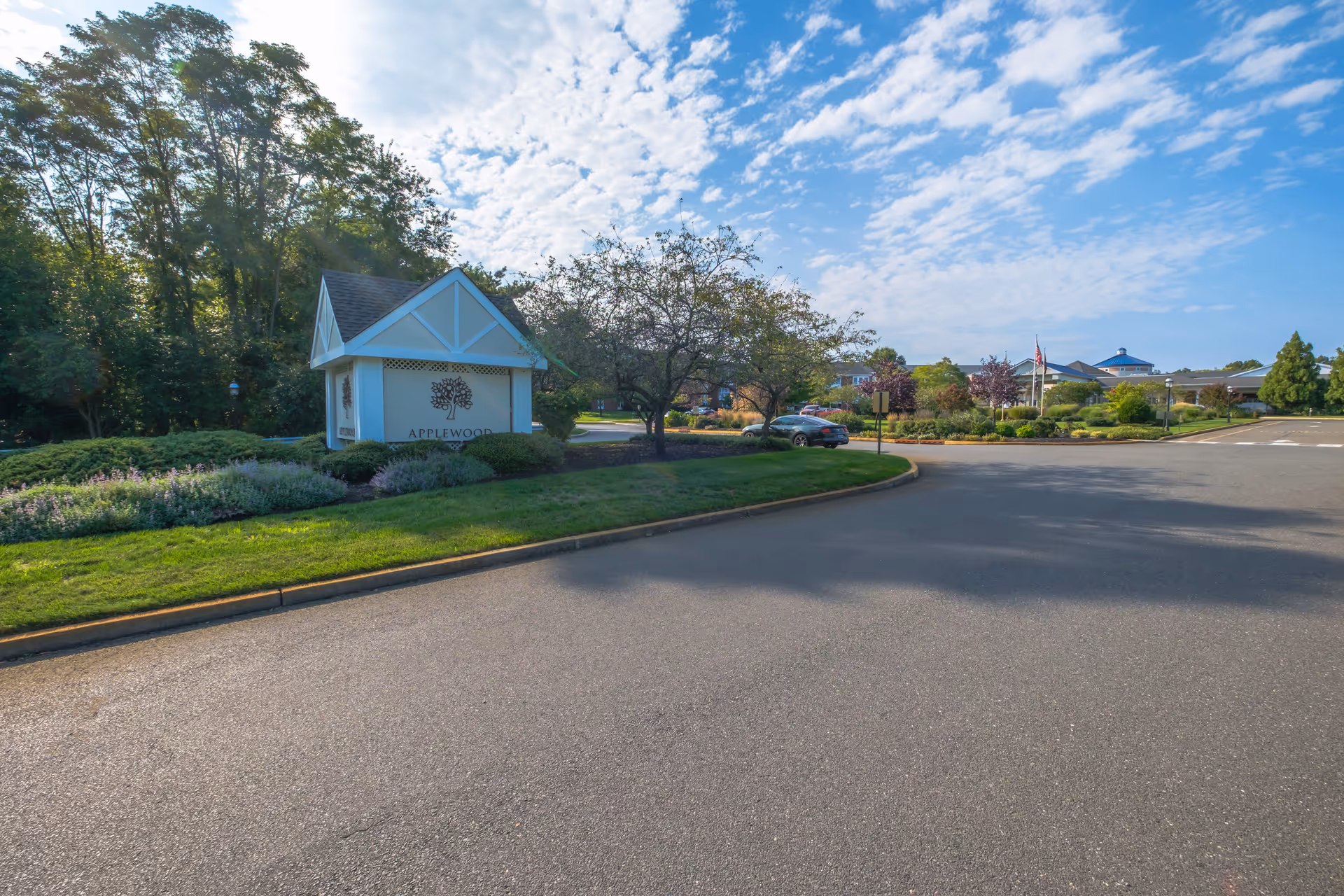 Entrance area of Applewood Village senior living facility with a landscaped sign featuring the facility's name and logo, surrounded by trees and bushes under a partly cloudy blue sky.