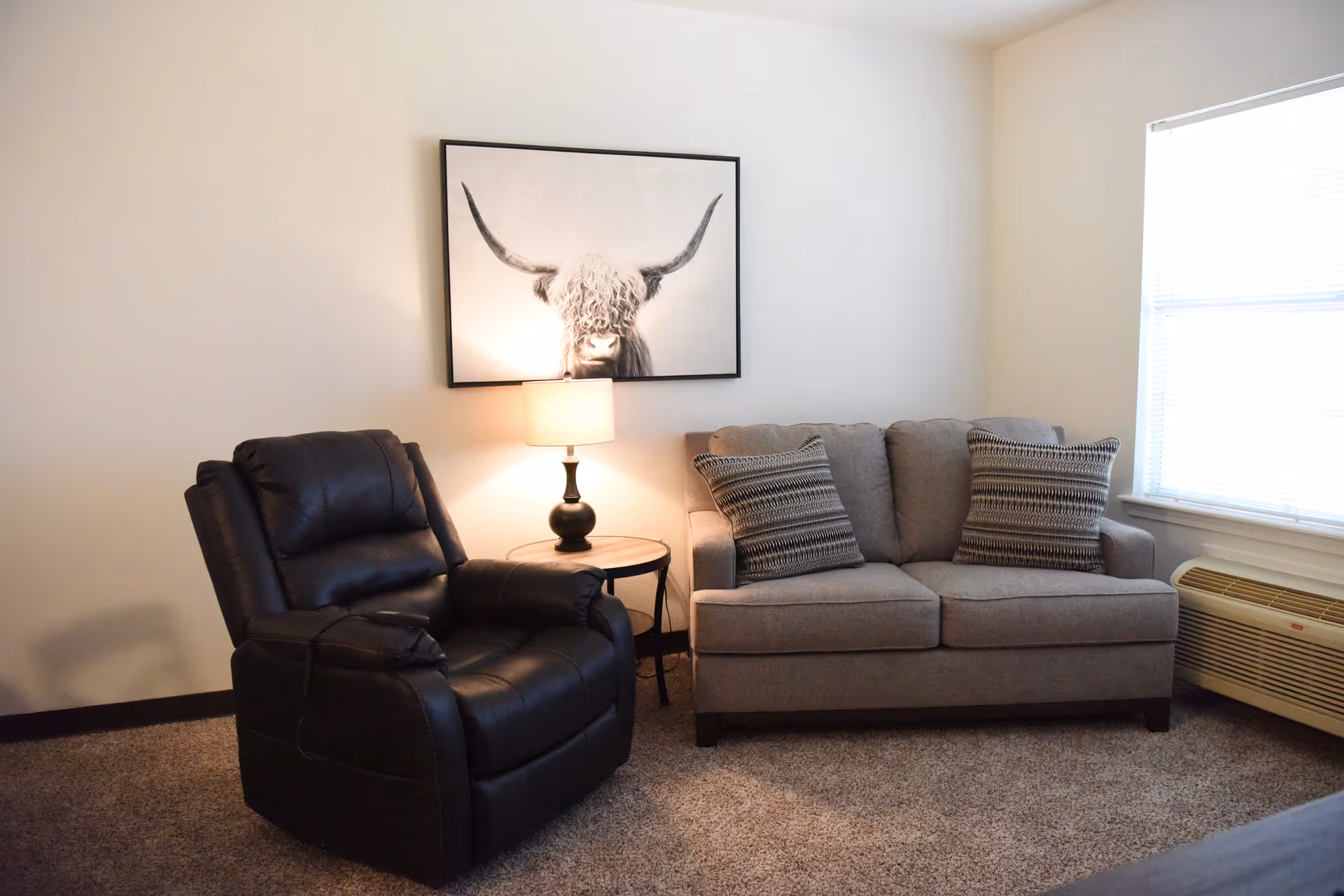 A cozy living room area with a black leather recliner chair and a gray fabric loveseat with two patterned pillows. Between them is a small round wooden side table with a lamp. Above the lamp hangs a framed black and white picture of a longhorn cow. A window with blinds and an air conditioning unit is on the right side of the room.