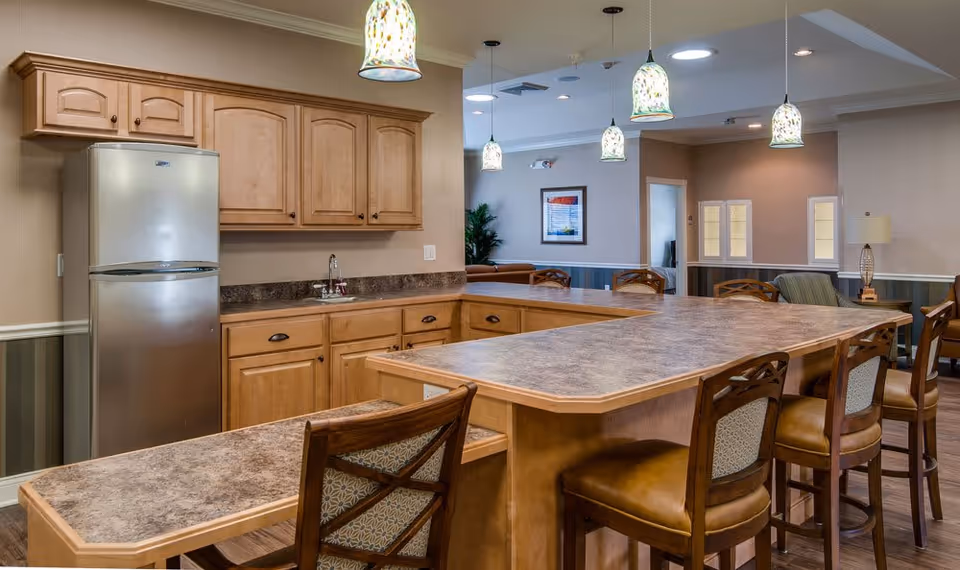 Interior view of a senior living facility kitchen and common area with wooden cabinets, a stainless steel refrigerator, a large countertop island with several chairs, and hanging pendant lights above. The background shows a seating area with framed artwork and soft lighting.