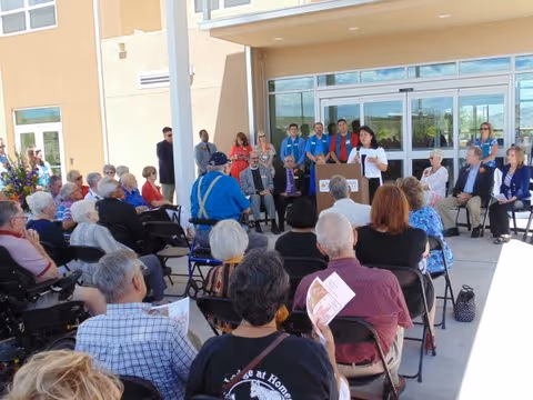 A group of elderly people seated outdoors in front of a building entrance, listening to a woman speaking at a podium. Several other people stand behind the speaker, and the audience is seated in folding chairs, some holding papers.
