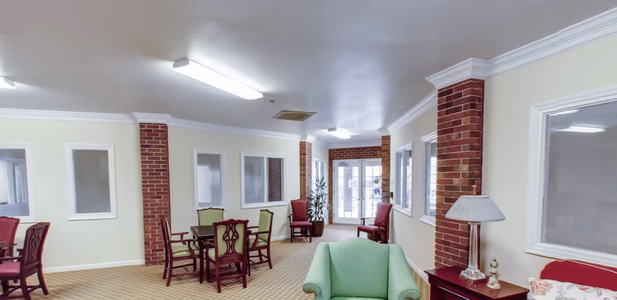 Bright senior living common room with dining tables and chairs, upholstered armchairs, brick columns, and a glass entrance.