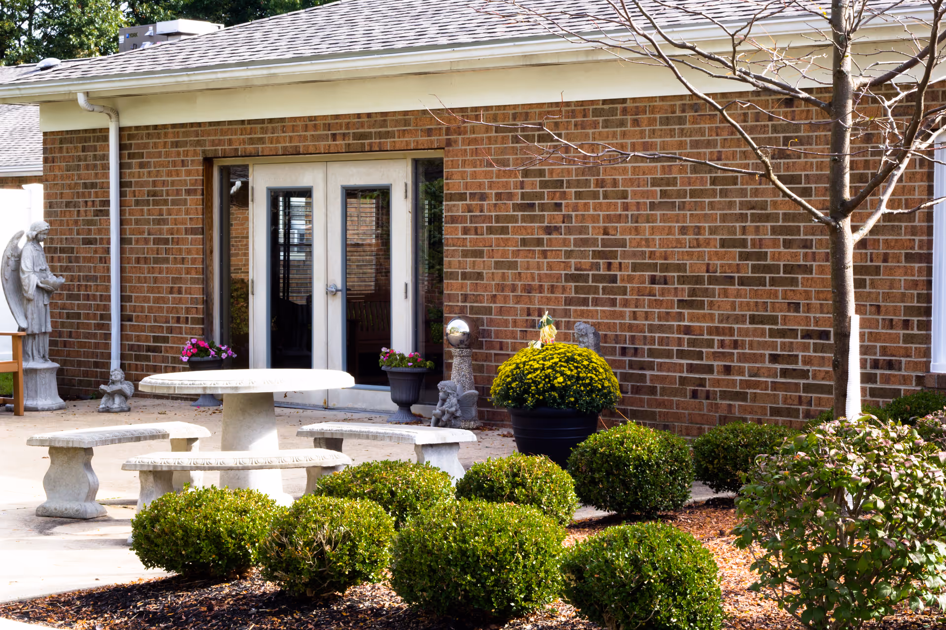 Outdoor patio area with a round stone table and matching stone benches surrounded by green bushes. There is a brick building with double glass doors in the background, along with potted plants and a stone angel statue on the left side.