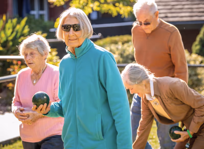 Four elderly people outdoors playing bocce ball on a sunny day, with greenery and a building in the background. One woman in a teal jacket and sunglasses is holding a bocce ball and looking at the camera.