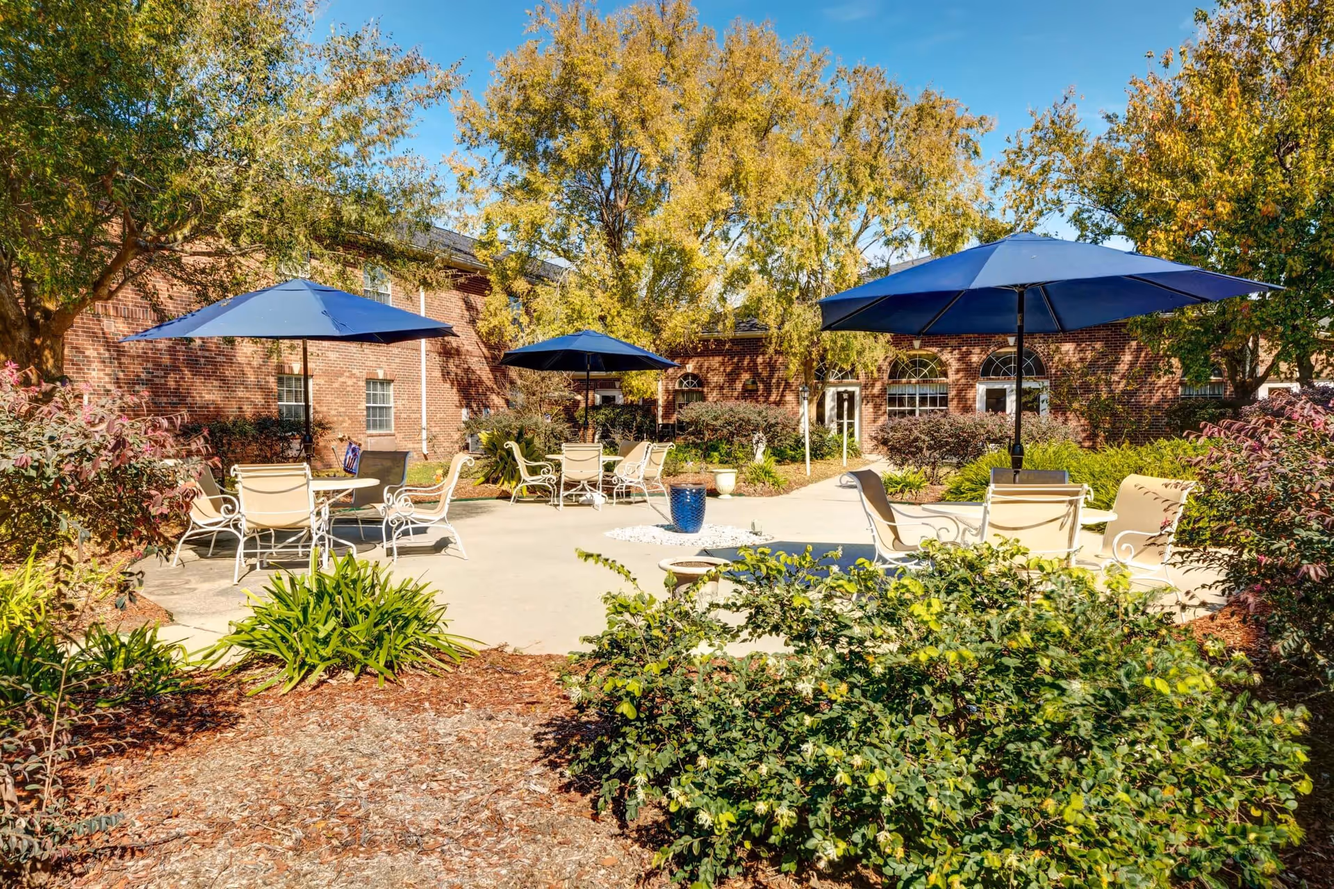 Outdoor patio area at Laketown Village with several round tables and chairs under large blue umbrellas, surrounded by green shrubs and trees with a brick building in the background under a clear blue sky.