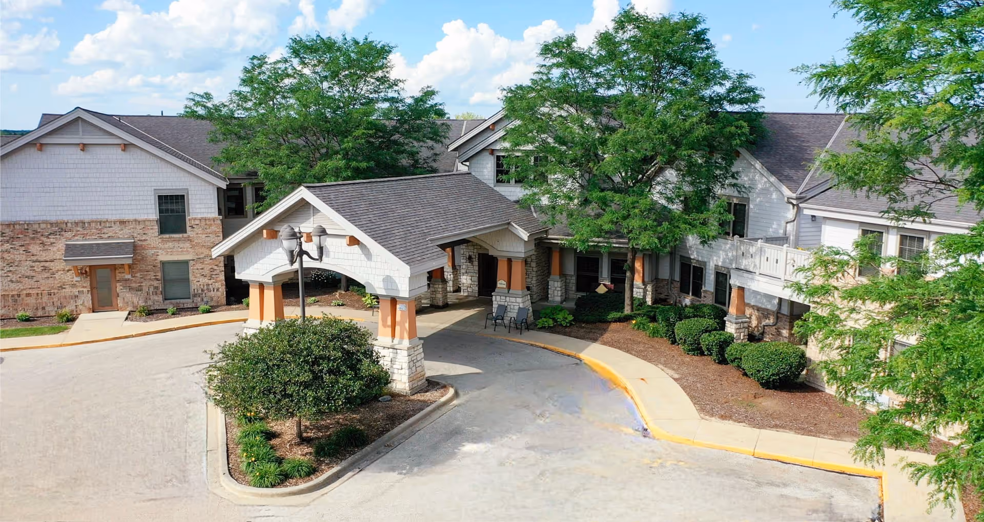 Exterior view of Riverview Village Senior Living facility showing a covered entrance with stone pillars and a driveway. The building has a combination of brick and white siding with multiple windows, surrounded by green trees and landscaped bushes under a partly cloudy sky.