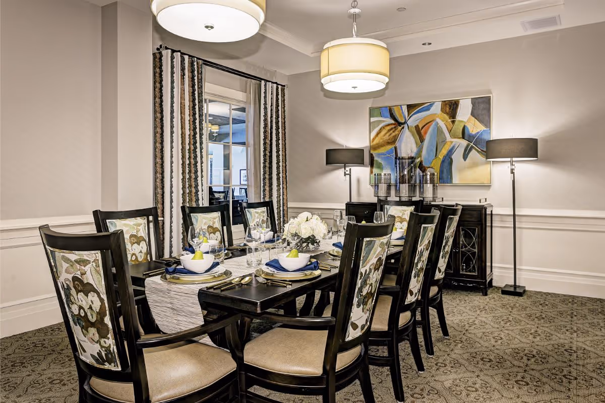 A well-decorated dining room with a long dark wooden table set for eight people. Each chair has a floral patterned backrest and beige seat cushion. The table is set with white bowls containing green apples, blue napkins, glassware, and silverware. The room features two large hanging ceiling lights, a floor lamp, a sideboard with decorative items, and a colorful abstract painting on the wall. There is a window with striped curtains on one side.
