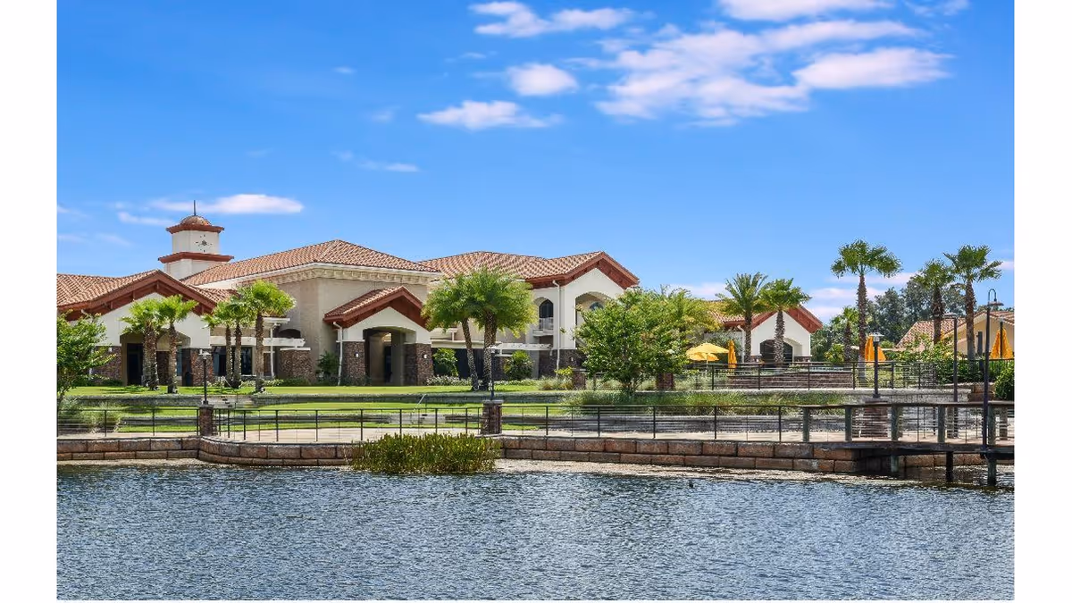 View of a large building with a tiled roof and multiple arches, surrounded by palm trees and greenery, situated next to a body of water with a stone and metal railing along the edge under a blue sky with some clouds.