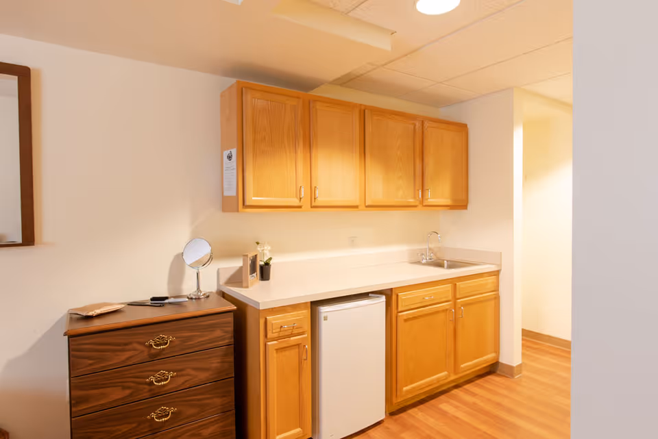Interior view of a small kitchenette area with wooden cabinets mounted on the wall and below the countertop. There is a small white refrigerator under the counter and a sink with a faucet on the right side. To the left of the kitchenette is a dark wooden dresser with three drawers, a small round mirror, and a few decorative items on top. The floor is wooden, and the walls are painted white.