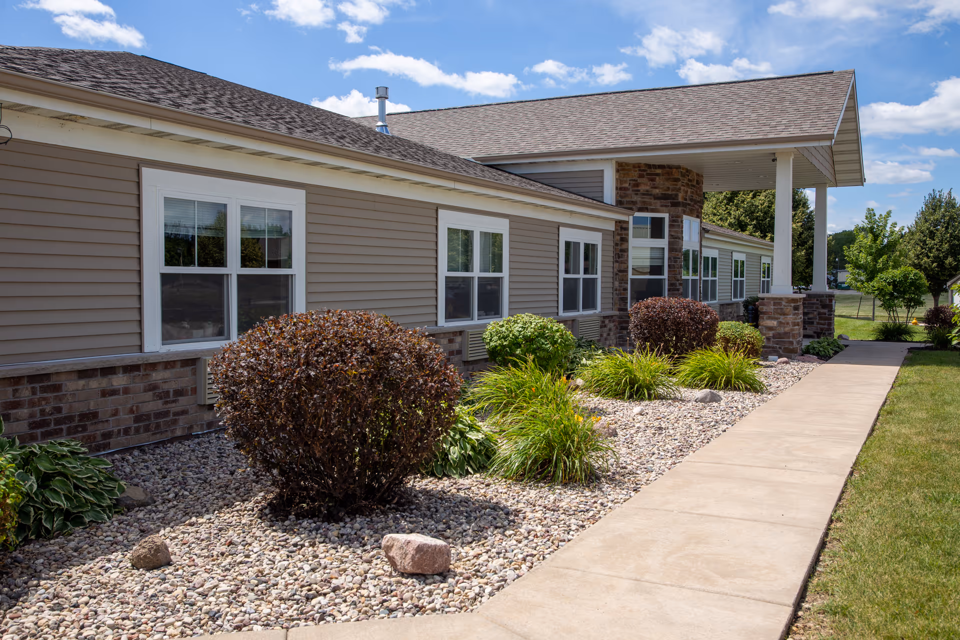 Exterior view of a single-story assisted living facility building with beige siding and stone accents. The building is surrounded by a landscaped garden with bushes, plants, and rocks, and a concrete walkway runs alongside the garden. The sky is blue with some clouds.