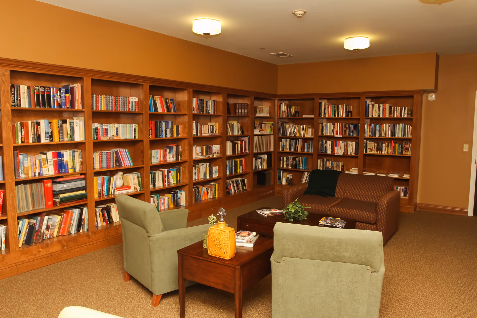 A cozy library room in an assisted living facility with wooden bookshelves filled with books lining the walls. The room has a brown couch, two green armchairs, a wooden coffee table with magazines and a decorative plant, and warm lighting from ceiling fixtures.