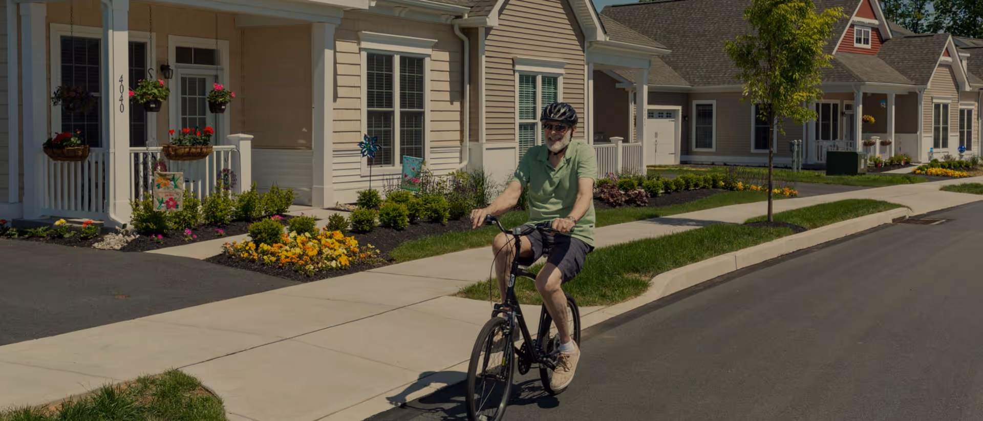 An elderly man wearing a helmet rides a bicycle on a paved street in front of a row of single-story residential buildings with porches, flower beds, and well-maintained landscaping on a sunny day.