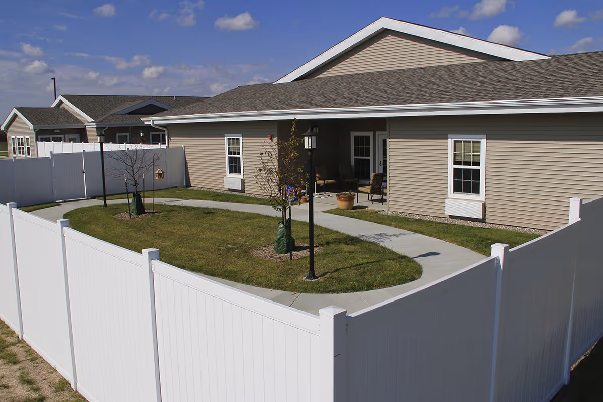 Outdoor courtyard area enclosed by a white fence with a concrete walkway, small trees, a lamp post, and a beige building with a covered patio and chairs under a blue sky with scattered clouds.