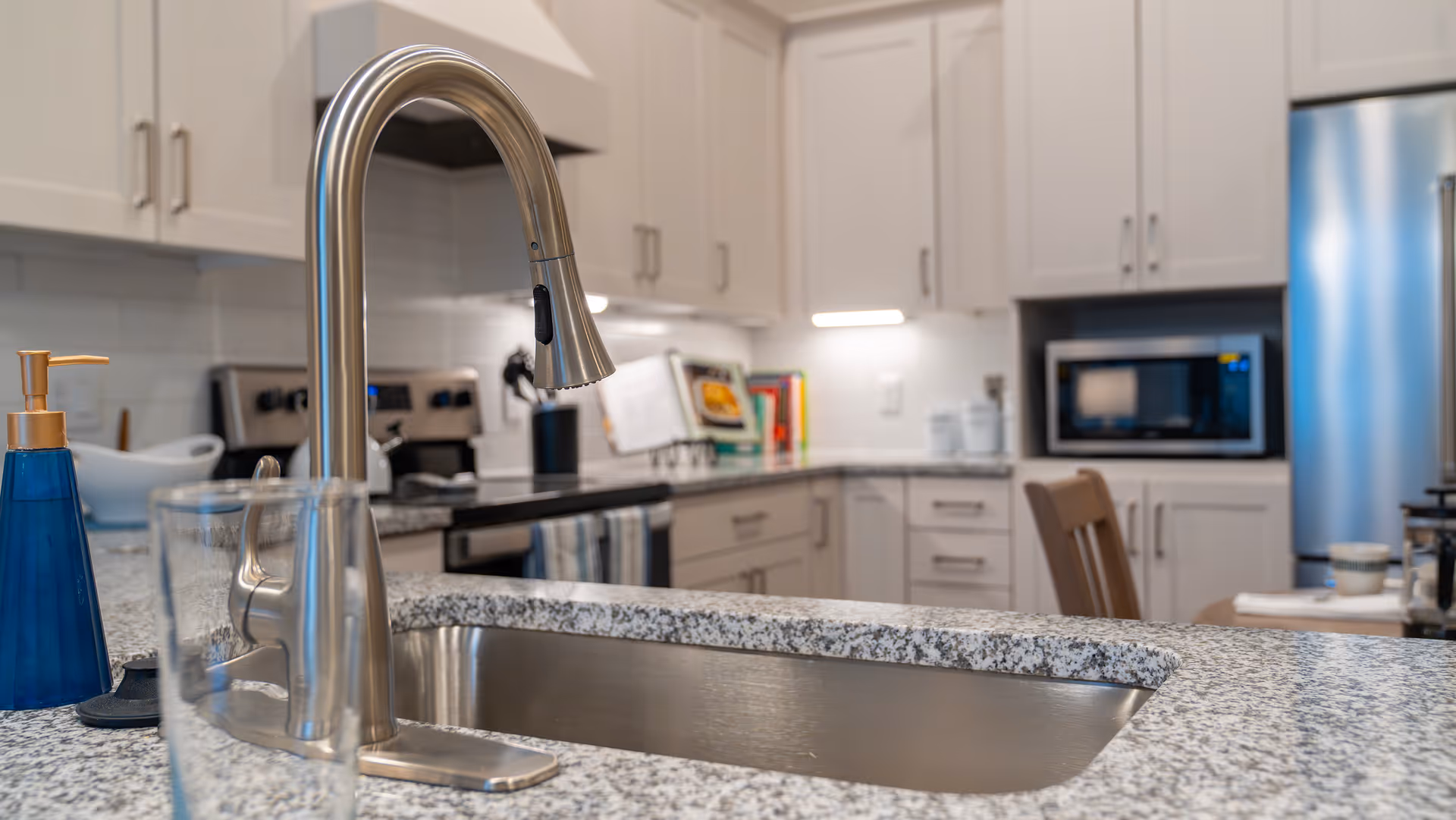 Modern kitchen sink and pull-down faucet on a granite countertop with white cabinets, microwave, and refrigerator in the background.