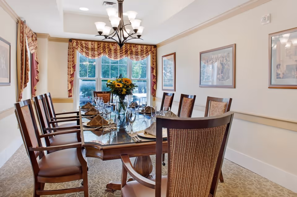 A formal dining room with a long wooden table set with plates, napkins, glasses, and silverware. The table has a centerpiece of sunflowers in a vase. The room features large windows with patterned curtains, beige walls adorned with framed pictures, and a chandelier hanging from the ceiling.