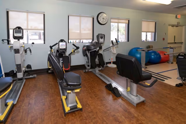 A fitness room with several exercise machines including recumbent bikes and elliptical trainers. The room has wooden flooring, three windows with blinds, a wall clock, and exercise balls in the corner near parallel bars.